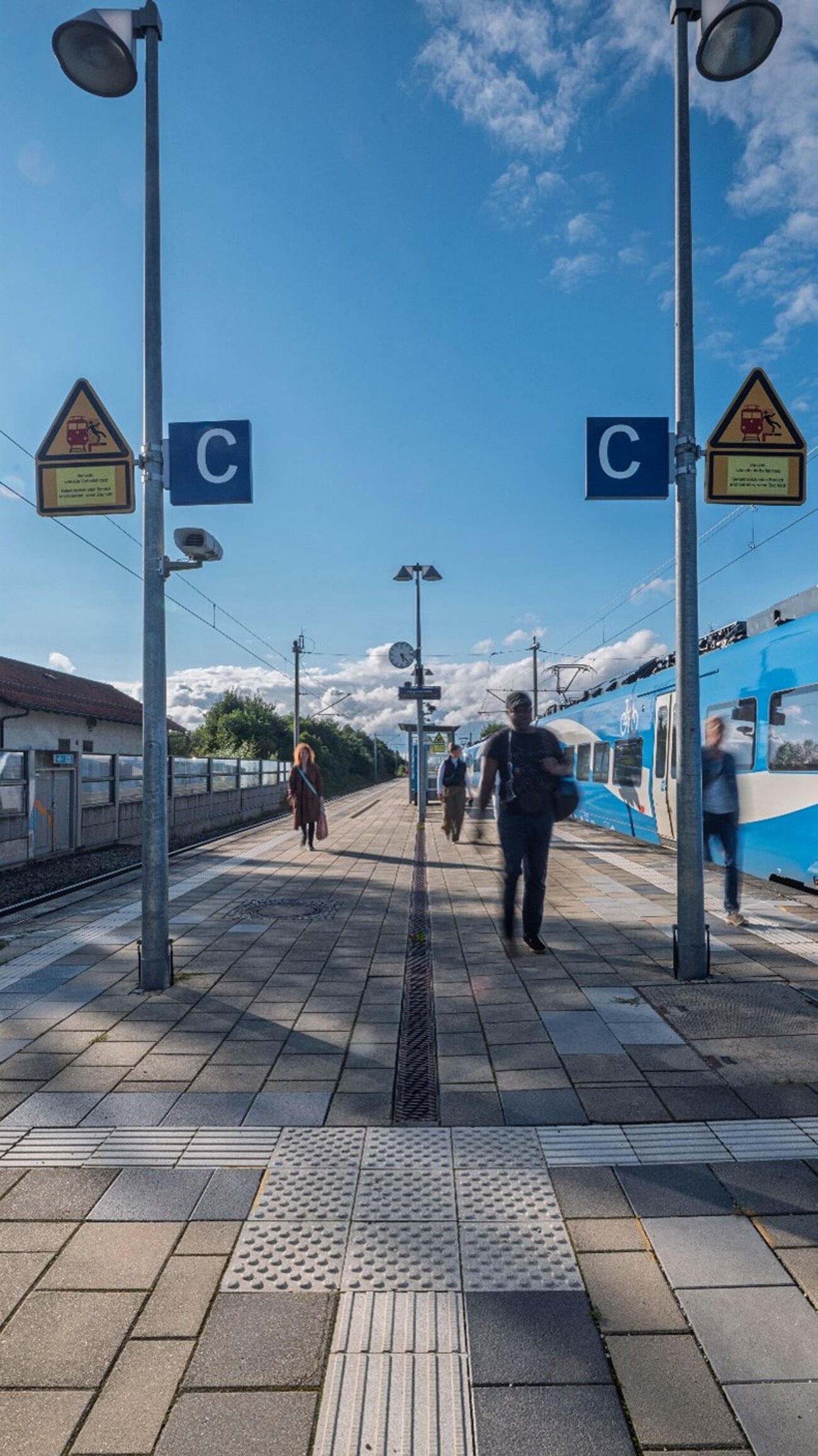 Farbfoto im Hochformat. Zu sehen ist ein Bahnsteig eines Bahn- oder S-Bahnhofs, rechts und links befinden sich die Gleise. Fahrgäste sind schemenhaft zu erkennen. Auf dem rechten Gleis ist ein blauer Zug zu sehen. 