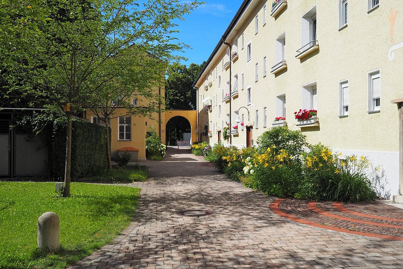 Farbfoto im Querformat, Es ist ein Innenhof zu sehen, der durch zwei dreigeschossige Häuserzeilen gebildet wird. Links befindet sich eine Rasenfläche mit Bäumen, der mittig zu sehende Weg ist gepflastert, an der rechts zu sehenden Häuserzeile wurden in Beeten Blumenstauden gepflanzt. Die Wohnanlage befindet sich in München Pasing. Es ist ein Beispiel für ein prämiertes Begrünungsprojekt.