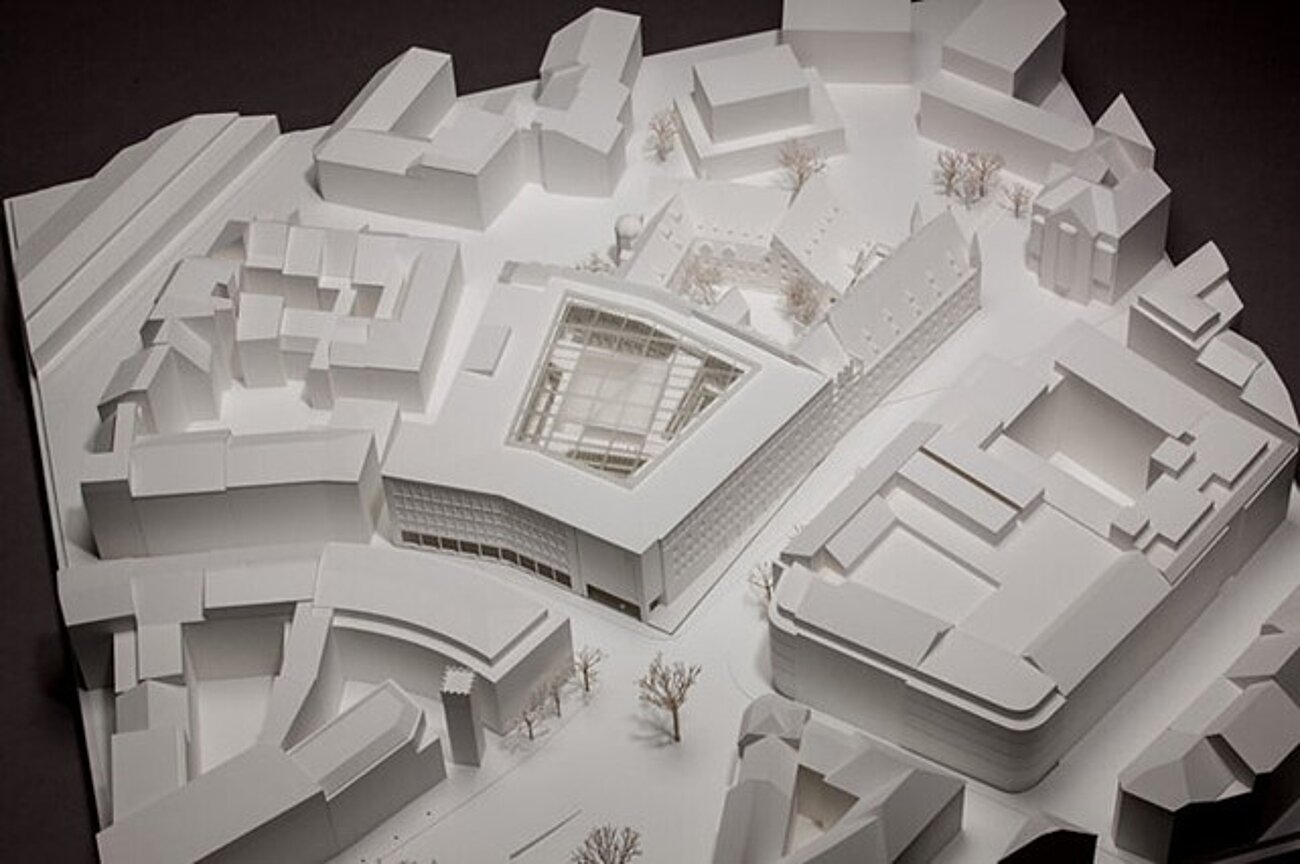 Farbfoto im Querformat. Das Foto zeigt das Architekturentwurfsmodell des zukünftigen Münchner Stadtmuseums und der anliegenden Gebäude, Straßen und Plätzen. Das Modell wurde aus weißen Kunststoffplatten gebaut. Zu erkennen ist der zukünftige Einbau in Form eines Kubus im Innenhof mit seinen Verstrebungen zu den Etagen des umliegenden Gebäudeteils. Auch das darüber liegende Glasdach ist zu sehen. 