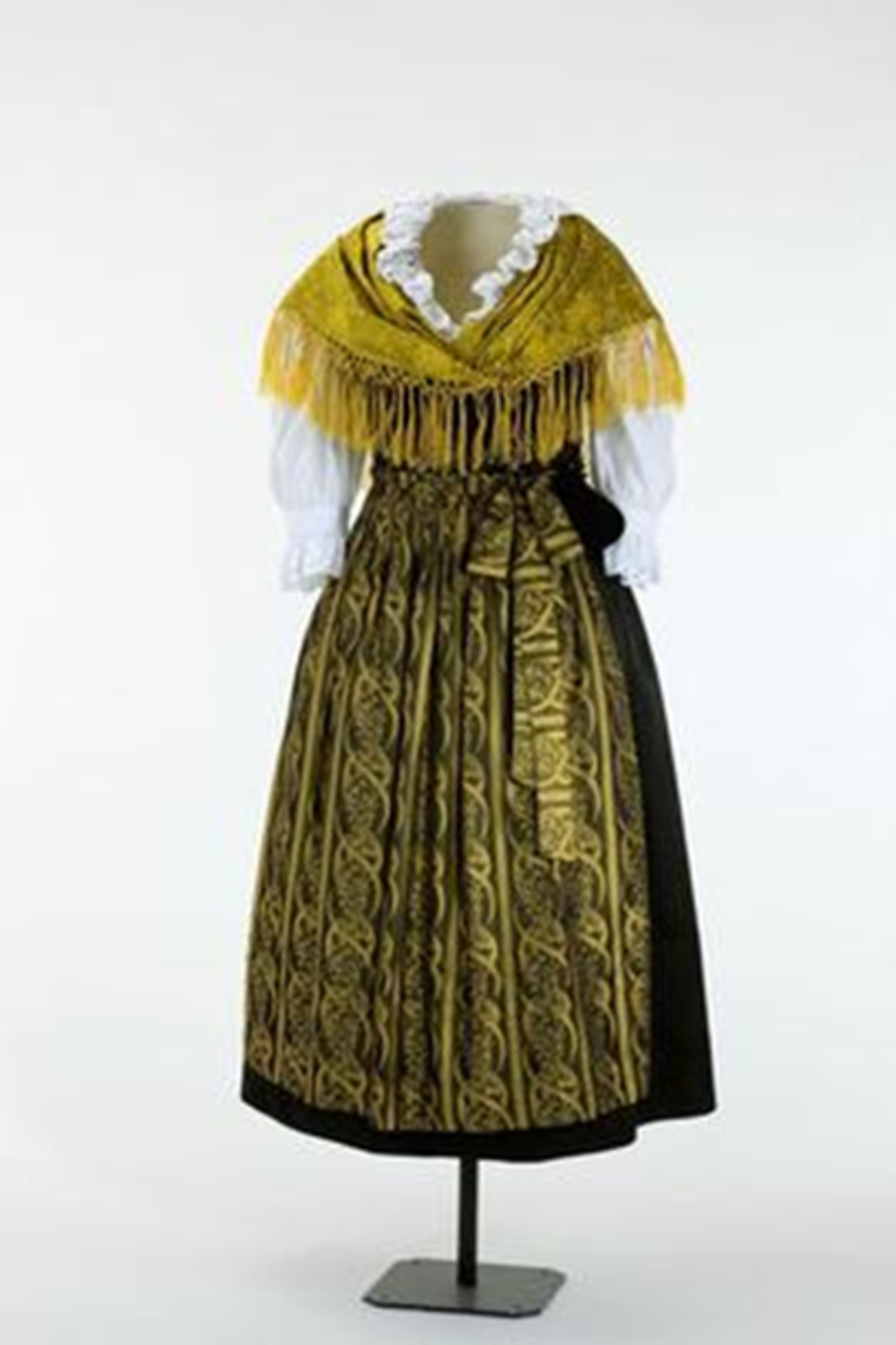 Farbfoto im Hochformat. Zu sehen ist ein Dirndlkleid auf einer Figurine vor hellem Hintergrund. Rock und Mieder sind aus einem aus dunklem Stoff genäht, dazu eine Schürze mit goldfarbenem Streifenmuster (Ornamente), eine weiße Dirndlbluse mit Spitzenbesatz sowie ein goldfarbenes gemustertes Schultertuch aus Damast mit Fransen. 