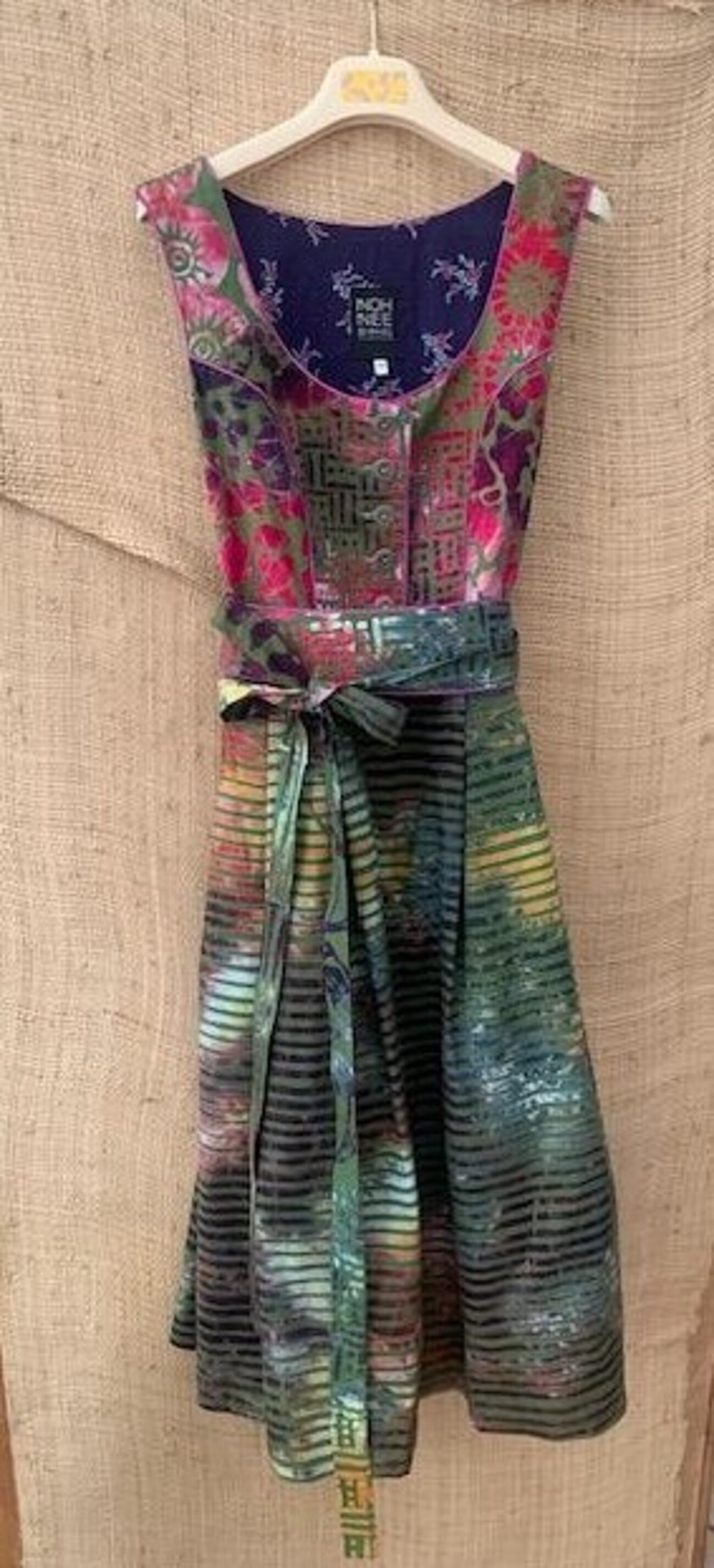 Farbfoto Hochformat. Mehrfarbiges in dunklen Grün- und Rot- und Gelbtönen gehaltenes Kleid im Stil eines Dirndls. Das Mieder ist mit Blumenornamenten versehen, der Rock mit Streifen und Ornamenten. Das Kleid hängt auf einem Kleiderbügel.