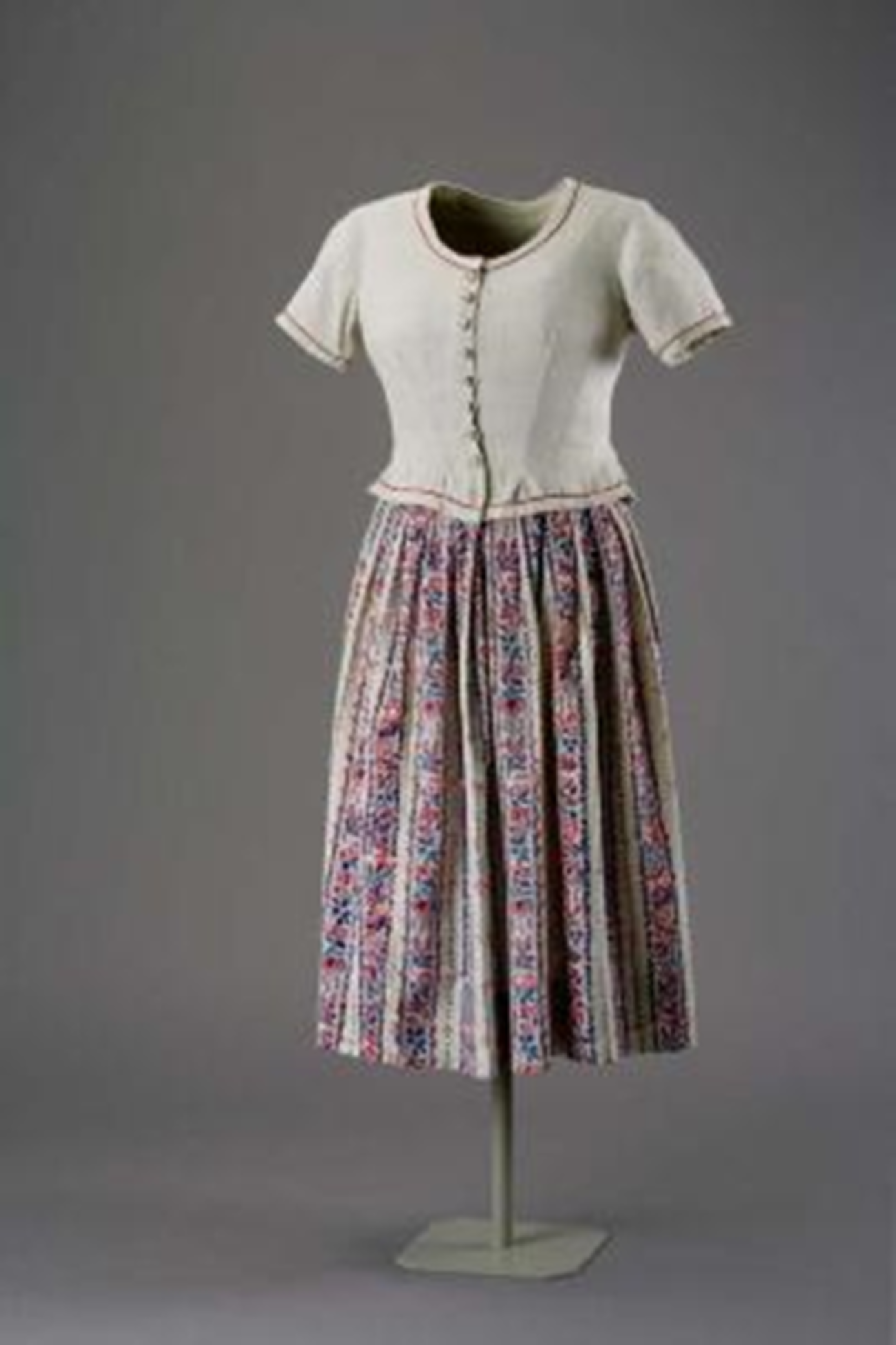 Farbfoto im Hochformat: Zu sehen ist ein Dirndlkleid auf einer Figurine vor dunklem Hintergrund. Das Kleid besteht aus einem Rock aus Baumwolle mit einem bäuerlichen Streifenmuster in Rot und Blau und einer naturfarbenen Spence-Jacke aus Leinen mit roten Baumwollpaspeln.