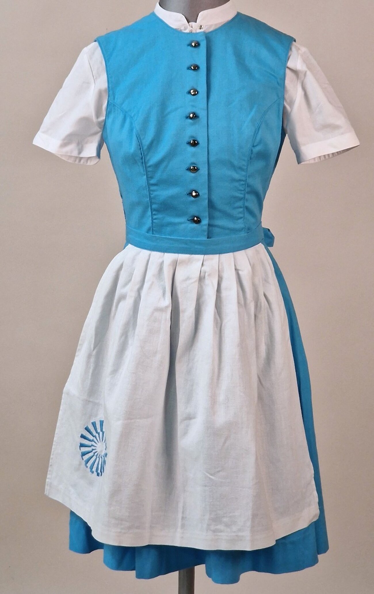 Farbfoto im Hochformat. Zu sehen ist ein Dirndlkleid auf einer Figurine vor hellem Hintergrund. Das Dirndlkleid besteht aus einem hellblauen kurzen Rock und einem hellblauen Mieder, einer weißen, kurzärmligen Bluse mit Stehkragen und einer weißen Schürze, auf der links ein blauer Strahlenkranz gestickt wurde. 