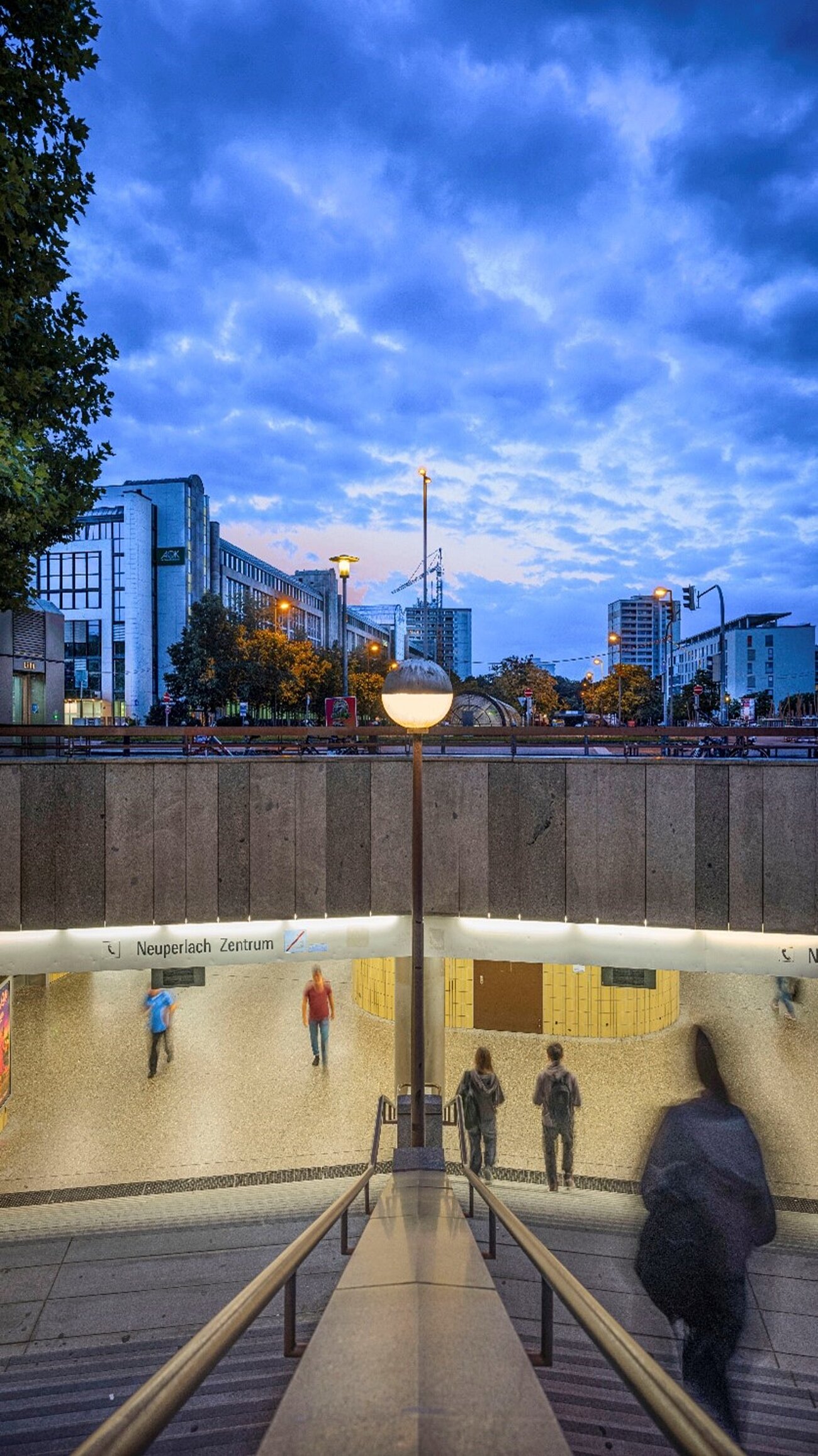 Farbfoto im Hochformat. In der unteren Bildhälfte ist ein zweigeteilter U- oder S-Bahn-Treppen Abgang zu sehen, der in das Zwischengeschoss eines unterirdischen Bahnhofs führt. Auf einem Hinweisschild ist „Neuperlach Zentrum“ zu lesen. Darüber kann man die dicke Betondecke erkennen. Im oberen Bildteil erheben sich Gebäude, Türme und eine Ampel in den dunkelblauen, dämmernden Himmel.