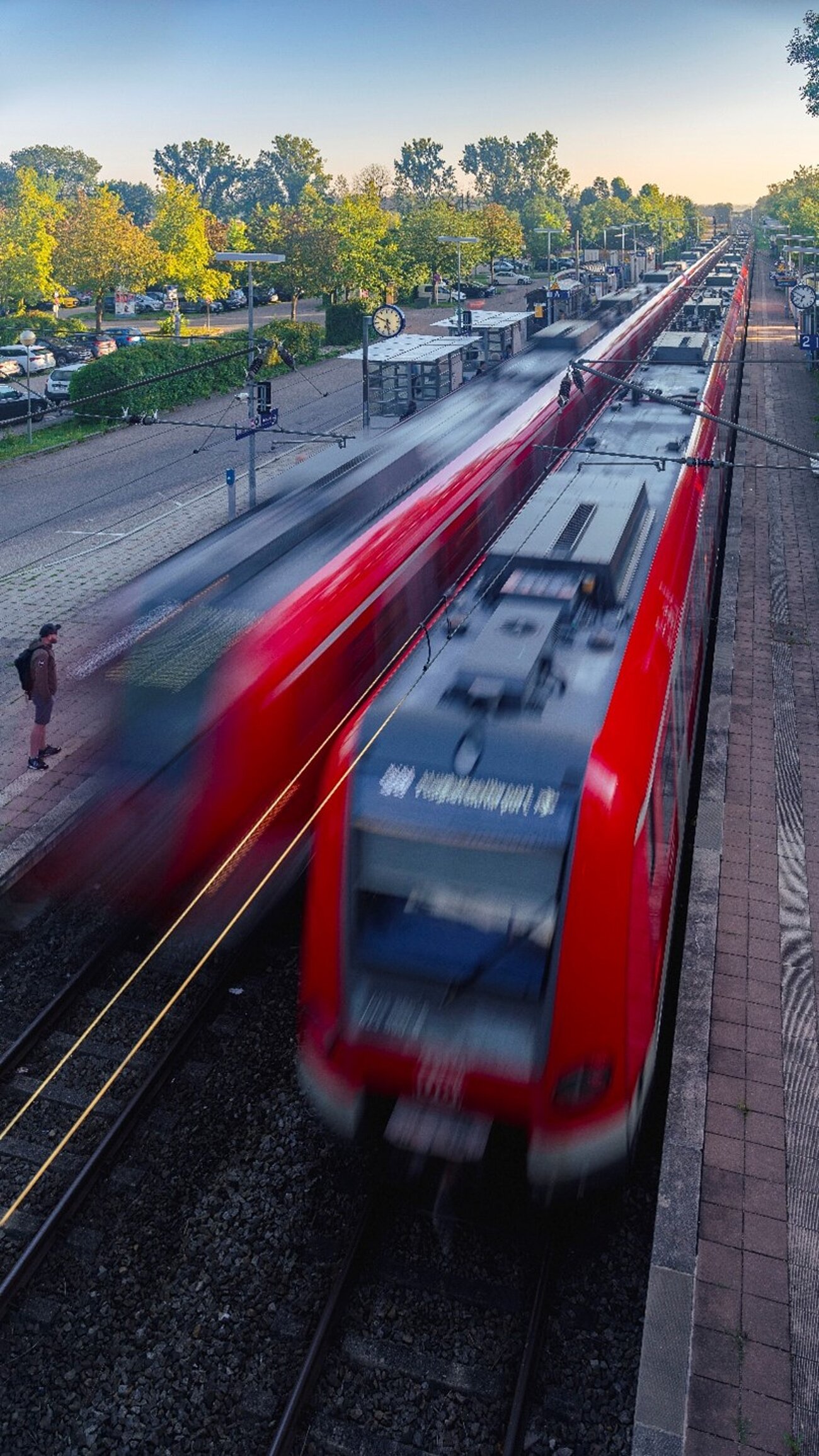 Farbfoto im Hochformat. Zu sehen ist ein Bahnsteig der S-Bahn mit einer einfahrenden und einer ausfahrenden S-Bahn. Am Gleisen stehen Fahrgäste. Im Hintergrund ist ein Parkplatz zu sehen, auf dem viele Auto parken.