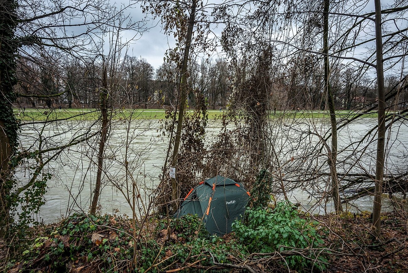 Farbfoto im Querformat. Zu sehen ist ein kleines dunkelgrünes Zelt im Gestrüpp am Isarufer. Die Isar fließt hinter dem Zelt entlang. Im Hintergrund ist der gegenüberliegende grüne Uferstreifen mit einem Weg zu erkennen. 