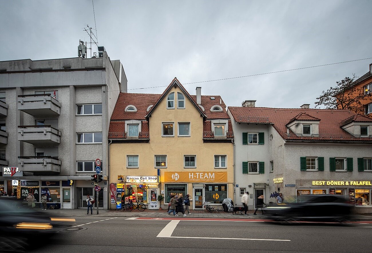 Farbfoto im Querformat. Häuserzeile in deren Mitte ein kleines gelbes Haus zu sehen ist, über dessen Ladenfenster ein großes Schild, beschriftet mit H-Team, montiert wurde. 