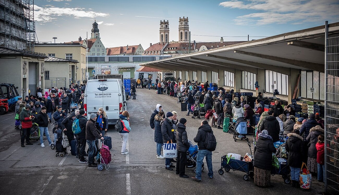 Farbfoto im Querformat. Auf einer Freifläche vor einer Hallenzeile mit Verladerampe zieht sich eine lange Schlange mit Menschen U-förmig über den asphaltierten Platz. Die Menschen haben Rolltaschen, Taschen, Rucksäcke u.ä. dabei. 
