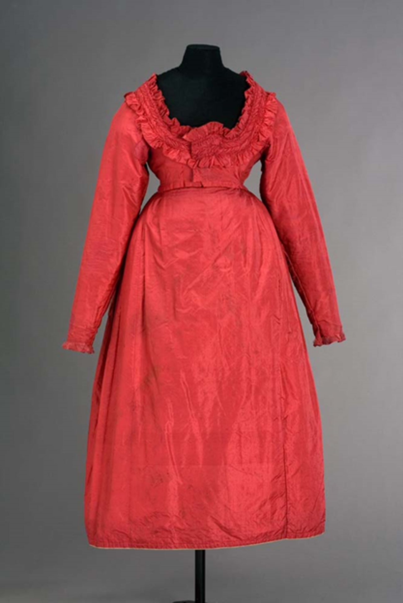 Farbfotografie, Hochformat. Zu sehen ist ein Dirndlkleid auf einer Figurine vor dunklem Hintergrund. Das Kleid wurde aus himbeerrotem Seidentaft genäht und besteht aus einem weiten bodenlangen Rock und einem Mieder mit Rüschen. 