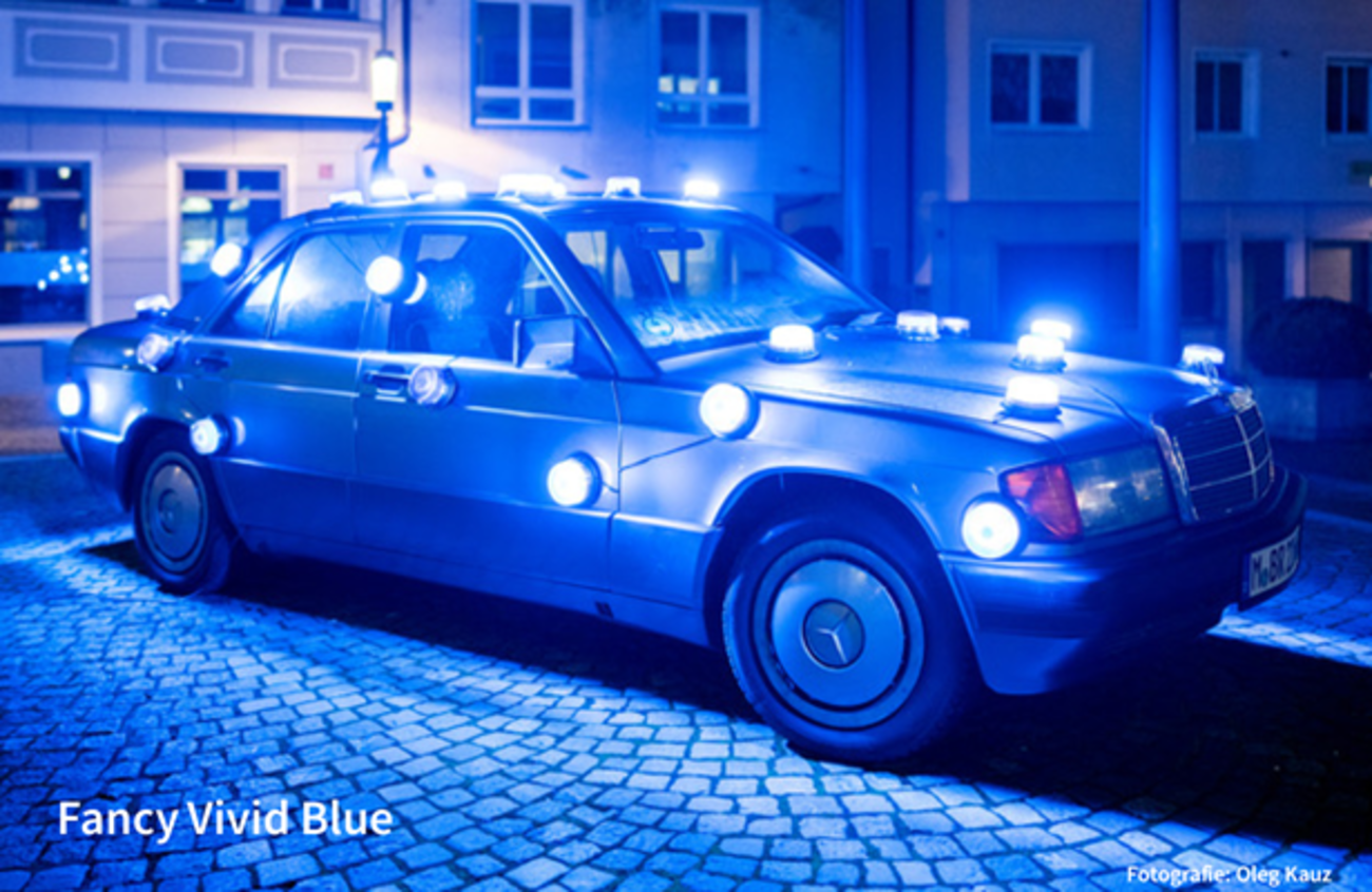 Farbfotografie im Querformat. Zu sehen ist ein alter, auf einem kleinen gepflasterten Platz stehender Mercedes Coupé. Es ist dunkel, der Platz ist durch eine Straßenlaterne etwas beleuchtet. Auf dem Dach und an der Seite des Autos sind runde Lampen angebracht, die leuchten, und Auto und Platz in ein bläuliches Licht tauchen. 